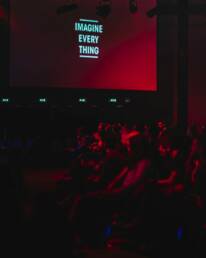 A dimly lit conference hall, with an audience watching a conference production with the words 'Imagine every thing' written on a slide in white letters.