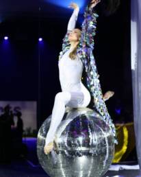 A lady dressed in white sat on a large disco ball with her arms in the air and a sequin trail coming from her arms, as GOTO Creates entertainment.