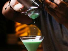 A close up of a bartender pouring a green cocktail into a martini glass as part of GOTO Creates Cocktail Masterclass