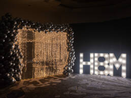 GOTO Creates Decor, dangling gold fairy light chains surrounded by black balloons. A sign reading HBM is lit up using white LED lights.