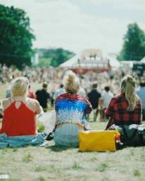 The back of three ladies sitting next to each other in a crowded field, with a blurred festival setup in the background.