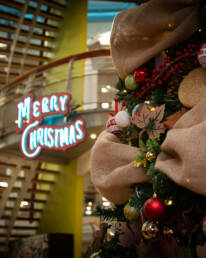 A close up of a Christmas tree in a multi-story building, with a lit-up merry Christmas sign on the staircase