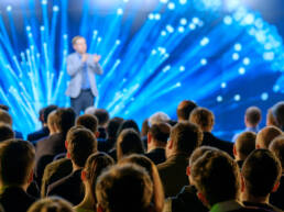 The audience listens to the lecturer at the conference hall. A screen of blue LEDs is behind him.