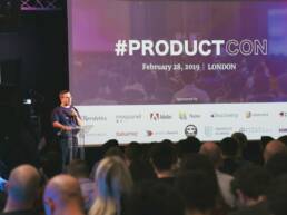 GOTO Creates Conference meeting, a speaker is talking with a microphone on stage and a screen is up with a slide titled #productcon